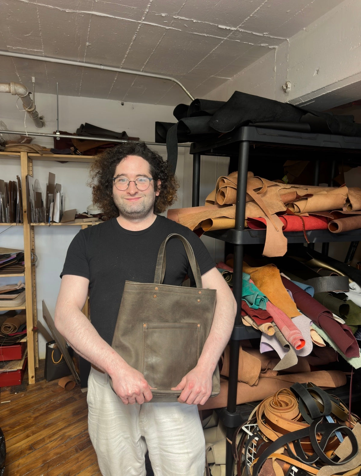 Man holding a leather bag in a workshop with leather pieces on shelves.